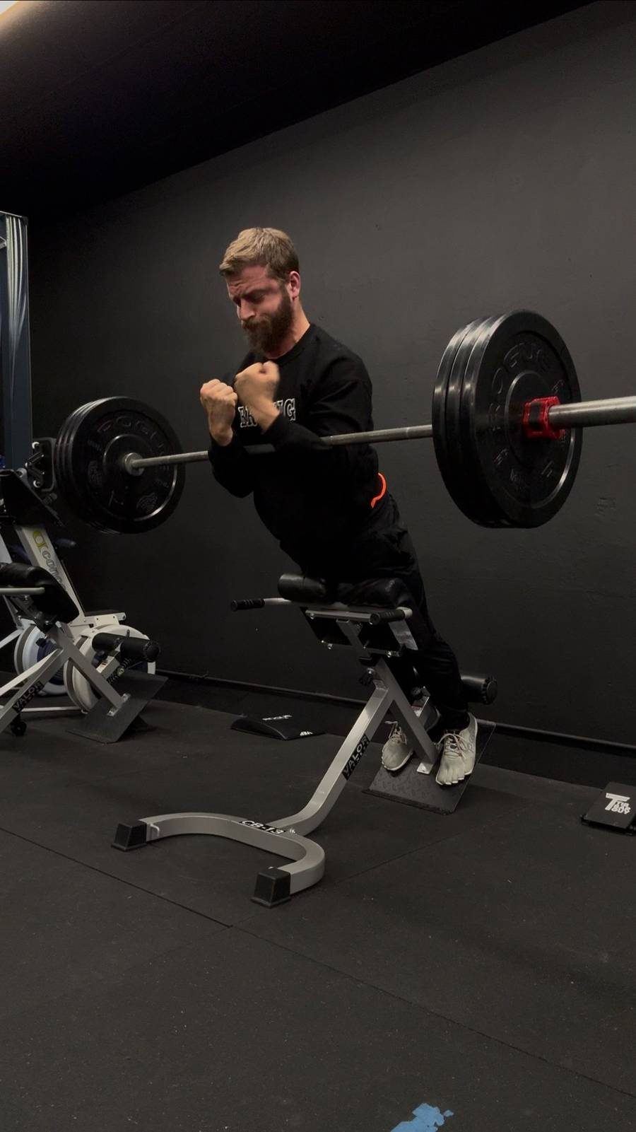 NWFIT coach and gym owner smiling while looking at phone in a dark gym, wearing a beige hoodie